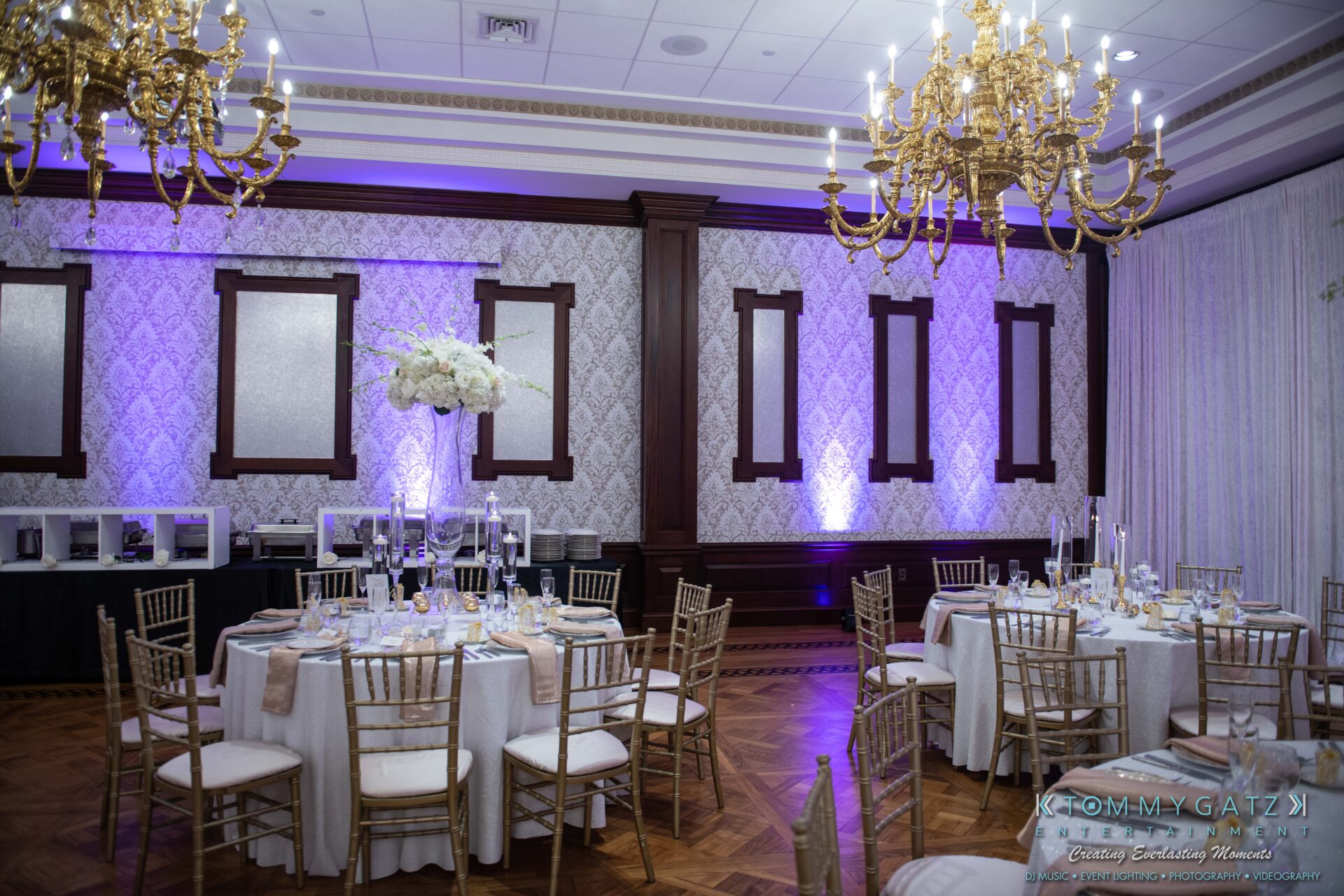 Harris-Wedding-HIGHLIGHTS-090119-87 - TOMMY GATZ ENTERTAINMENT Elegant banquet hall with round tables, white tablecloths, gold chairs, and floral centerpieces. Chandeliers hang from the ceiling, and purple lighting accents the patterned walls.