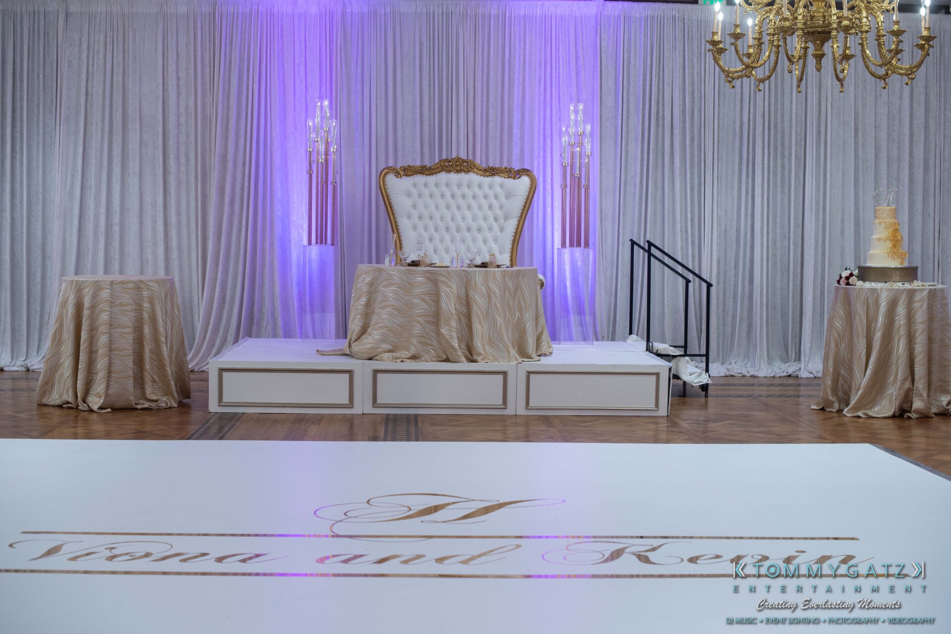 Harris-Wedding-HIGHLIGHTS-090119-88 - TOMMY GATZ ENTERTAINMENT A luxurious wedding reception setup with a grand white chair on a draped stage, chandeliers, and a monogrammed dance floor. Elegant drapery and soft lighting complete the scene.
