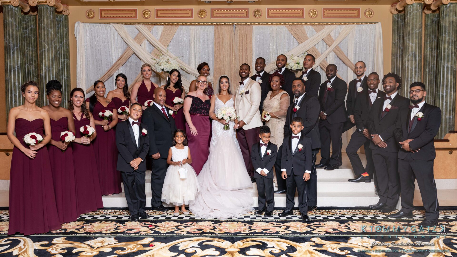 Harris-Wedding-HIGHLIGHTS-090119-89 - TOMMY GATZ ENTERTAINMENT A large wedding party poses together on a decorated stage. The group includes people in formal dresses and suits, holding bouquets.
