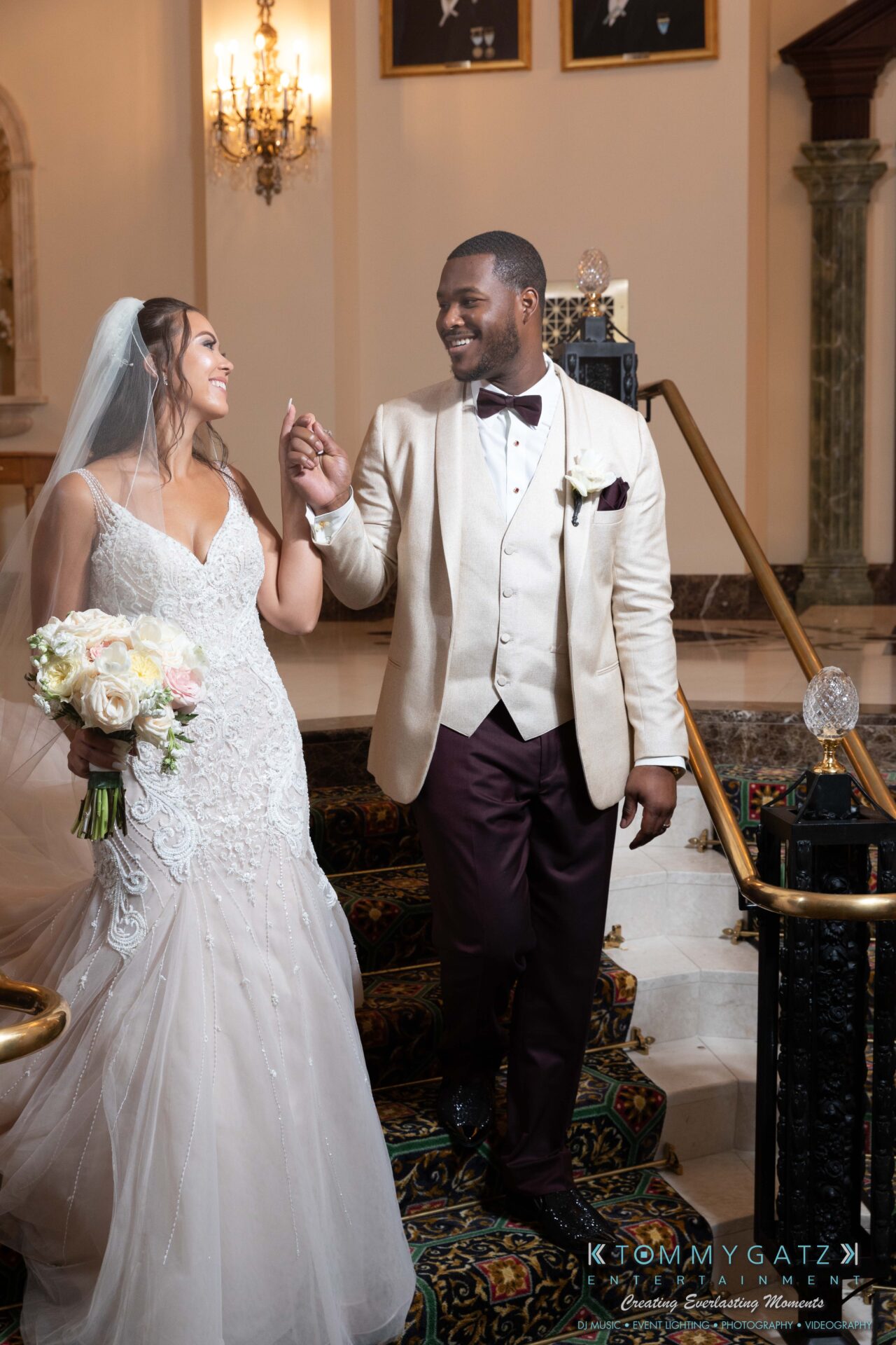 Harris-Wedding-HIGHLIGHTS-090119-92 - TOMMY GATZ ENTERTAINMENT A bride and groom in formal attire walk down a staircase, holding hands and smiling. The bride carries a bouquet of flowers.