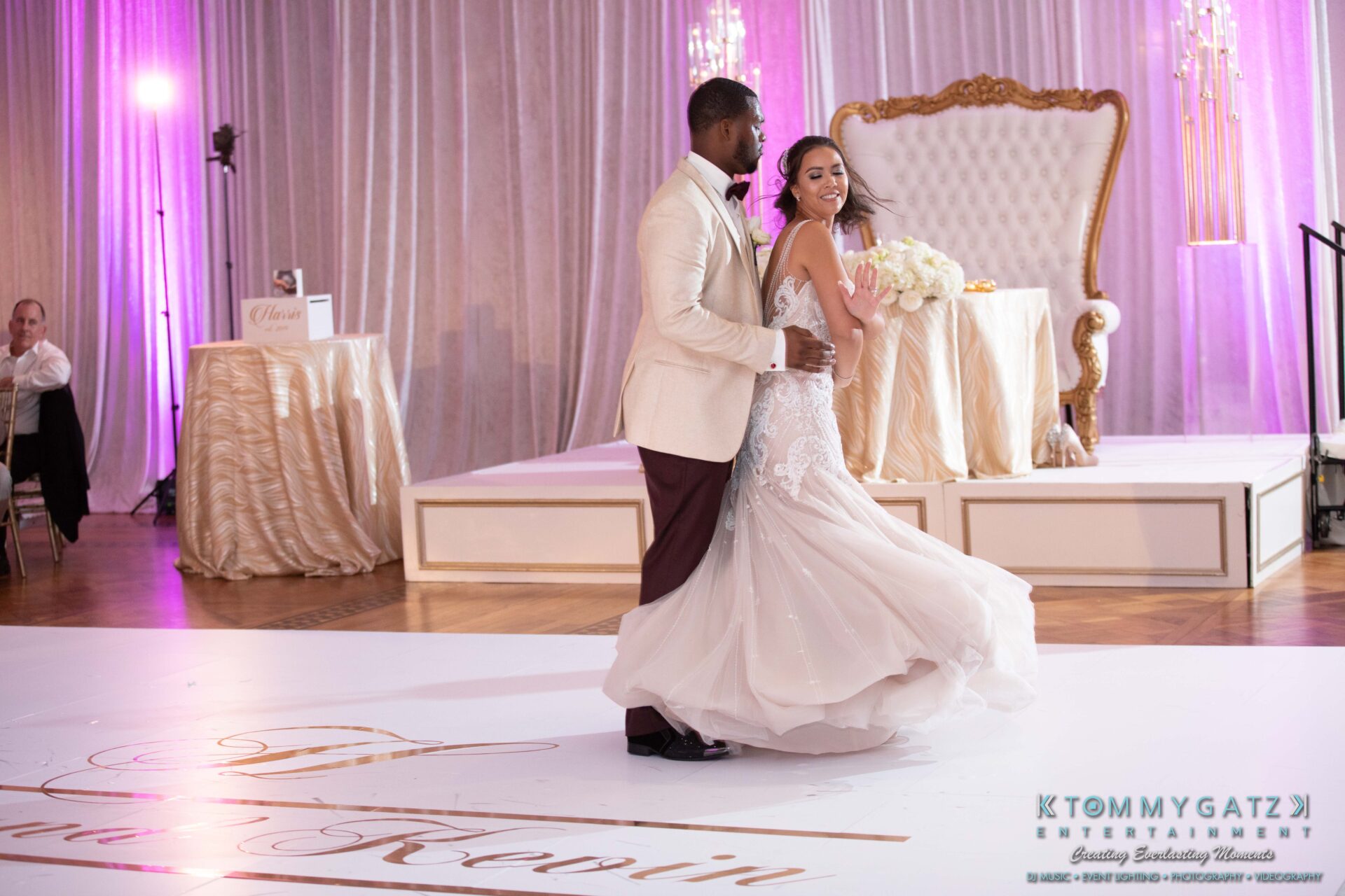 Harris-Wedding-HIGHLIGHTS-090119-94 - TOMMY GATZ ENTERTAINMENT A couple dances on a decorated ballroom floor, with a large white and gold chair and a flower arrangement in the background.