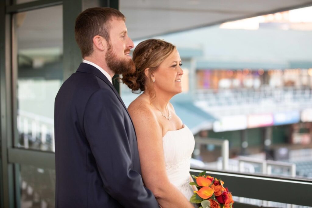 Heather-Scott-Wedding-Low-Res-100 1 - TOMMY GATZ ENTERTAINMENT A couple in wedding attire stands on a balcony overlooking a stadium. The bride holds a bouquet of orange flowers.