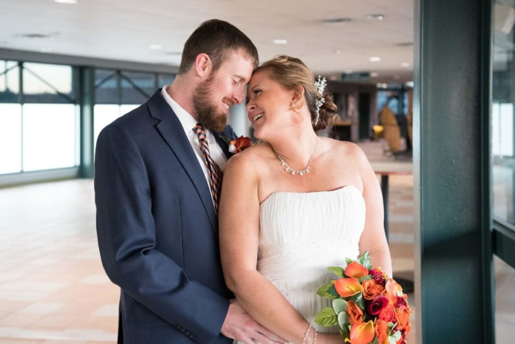 Heather-Scott-Wedding-Low-Res-101 1 - TOMMY GATZ ENTERTAINMENT A bride and groom share a moment indoors, smiling and leaning towards each other. The bride holds a bouquet of orange and red flowers.