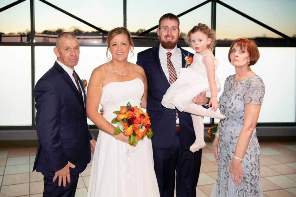 Heather-Scott-Wedding-Low-Res-103 1 - TOMMY GATZ ENTERTAINMENT A wedding portrait with five people. A bride in a white dress holds a bouquet, standing beside a groom in a suit who is holding a child. Two older adults stand on either side, all posed indoors.