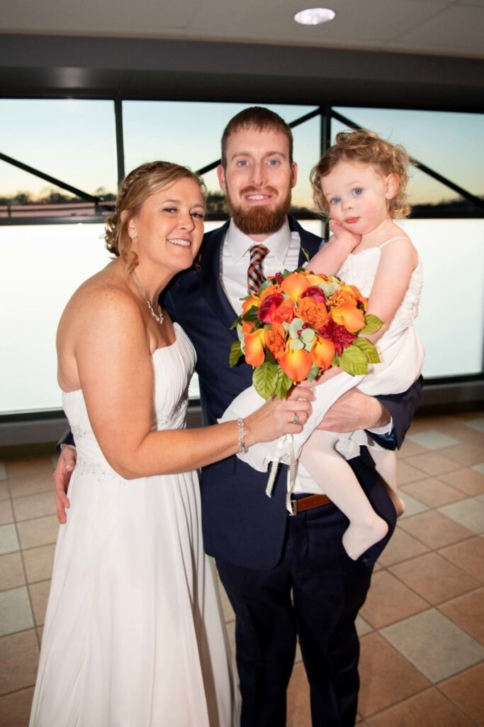Heather-Scott-Wedding-Low-Res-105 1 - TOMMY GATZ ENTERTAINMENT A smiling couple in formal attire poses with a young girl inside a building. The woman holds a colorful bouquet.