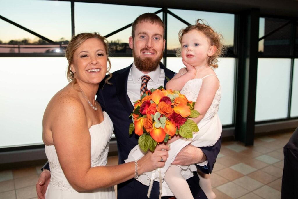 Heather-Scott-Wedding-Low-Res-106 1 - TOMMY GATZ ENTERTAINMENT A couple in wedding attire poses indoors with a young child. The bride holds a bouquet of orange and red flowers.