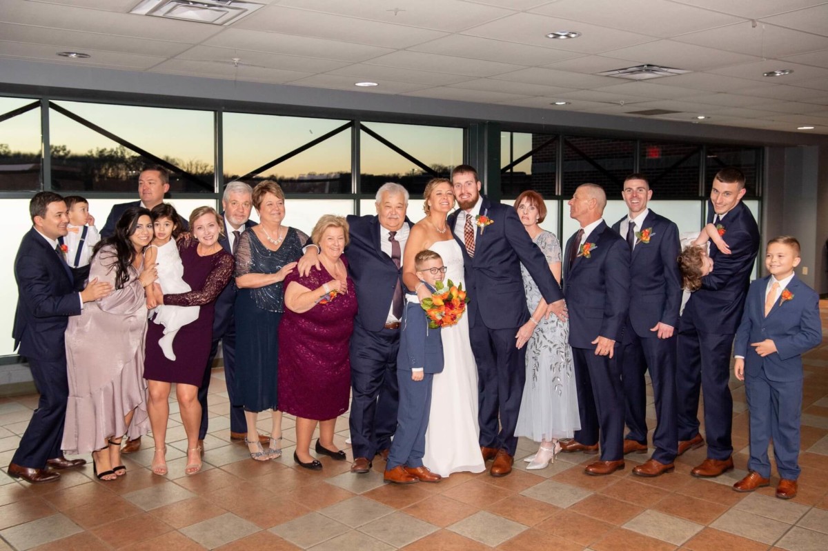 Heather-Scott-Wedding-Low-Res-107 1 - TOMMY GATZ ENTERTAINMENT A group of people, including a bride and groom, pose indoors for a photo. They are dressed in formal attire, with some holding bouquets. The setting appears to be a reception area.