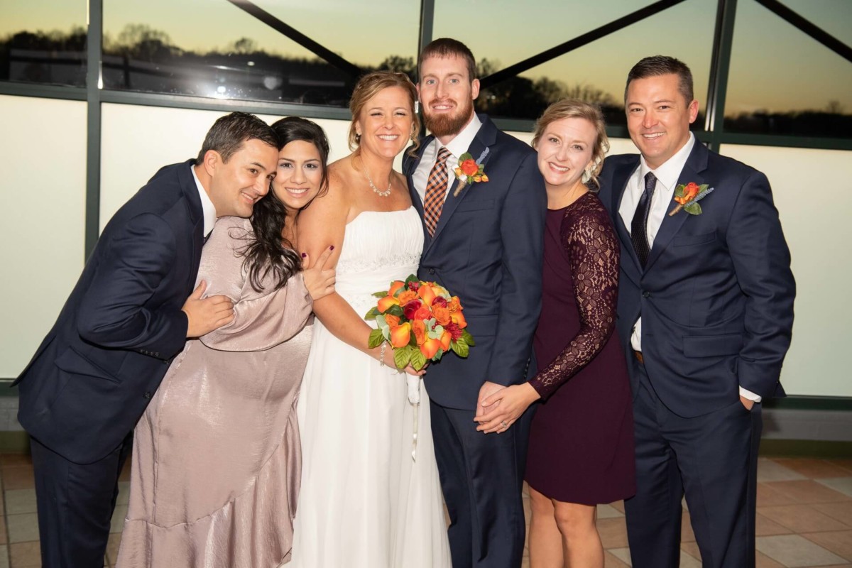 Heather-Scott-Wedding-Low-Res-108 1 - TOMMY GATZ ENTERTAINMENT A group photo of six people dressed formally, with a bride in the center holding a bouquet, standing in front of a large window at sunset.