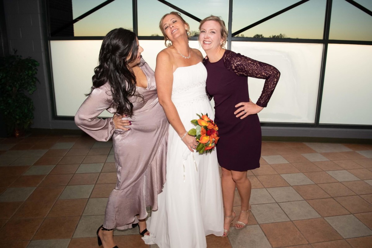 Heather-Scott-Wedding-Low-Res-109 1 - TOMMY GATZ ENTERTAINMENT Three women posing indoors, with the one in the center wearing a bridal gown and holding a bouquet. The others are dressed in purple and silver dresses. They are smiling and standing on a tiled floor.