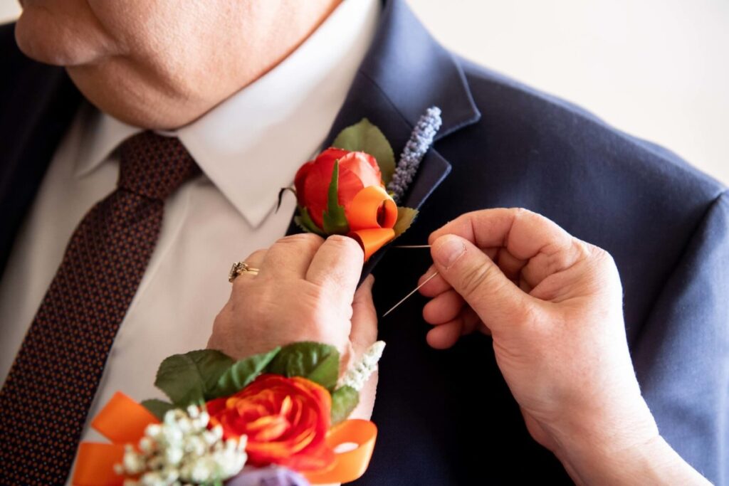 Heather-Scott-Wedding-Low-Res-11 1 - TOMMY GATZ ENTERTAINMENT Hands pin a red boutonniere on a man's dark suit jacket, with a red rose corsage visible on the wrist.