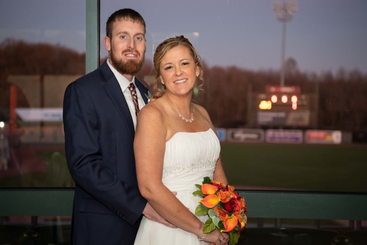 Heather-Scott-Wedding-Low-Res-110 1 - TOMMY GATZ ENTERTAINMENT A couple poses together in wedding attire, with the groom in a navy suit and the bride in a white dress holding a bouquet. A stadium is visible in the background through the window.