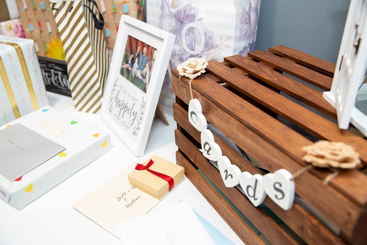 Heather-Scott-Wedding-Low-Res-111 1 - TOMMY GATZ ENTERTAINMENT Table with gifts and cards, featuring a wooden box labeled "cards" with heart-shaped tags, various wrapped presents, and a framed photo in the background.