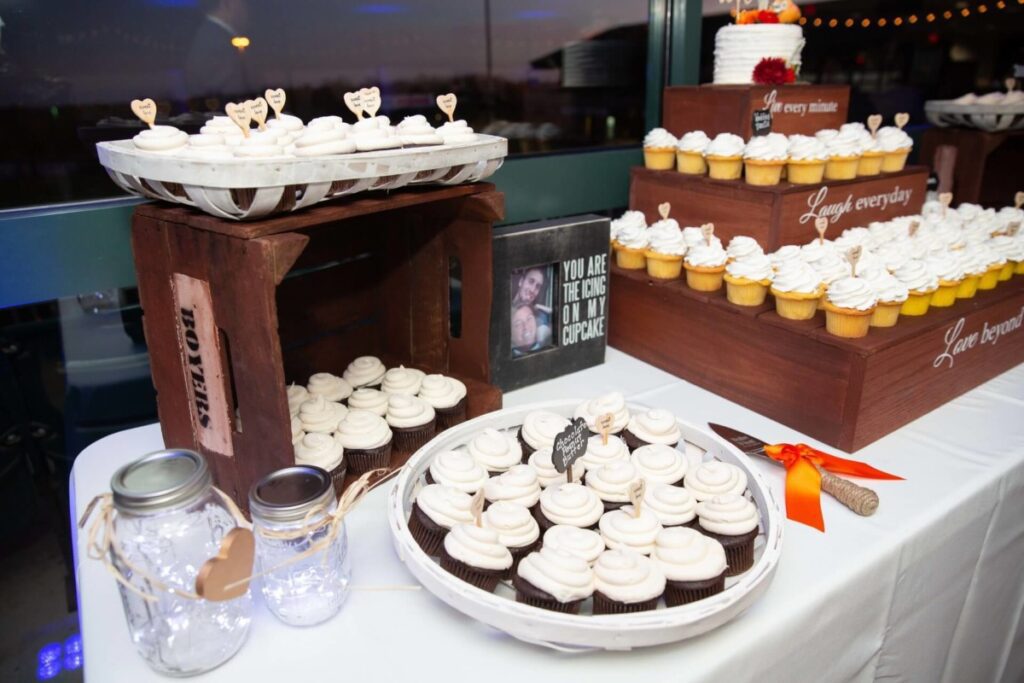 Heather-Scott-Wedding-Low-Res-112 1 - TOMMY GATZ ENTERTAINMENT A dessert table with white frosted cupcakes on white plates and wooden stands. Mason jars are in the foreground, and a framed sign is displayed among the cupcakes.