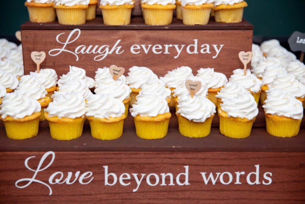 Heather-Scott-Wedding-Low-Res-113 1 - TOMMY GATZ ENTERTAINMENT A display of yellow cupcakes with white frosting on a wooden stand featuring the phrases "Laugh everyday" and "Love beyond words" in white lettering.