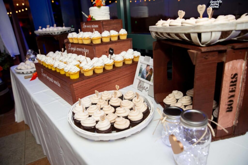 Heather-Scott-Wedding-Low-Res-114 1 - TOMMY GATZ ENTERTAINMENT Display of cupcakes on wooden crates at an event with decorative lights and photo frame; mason jars with string lights in front.