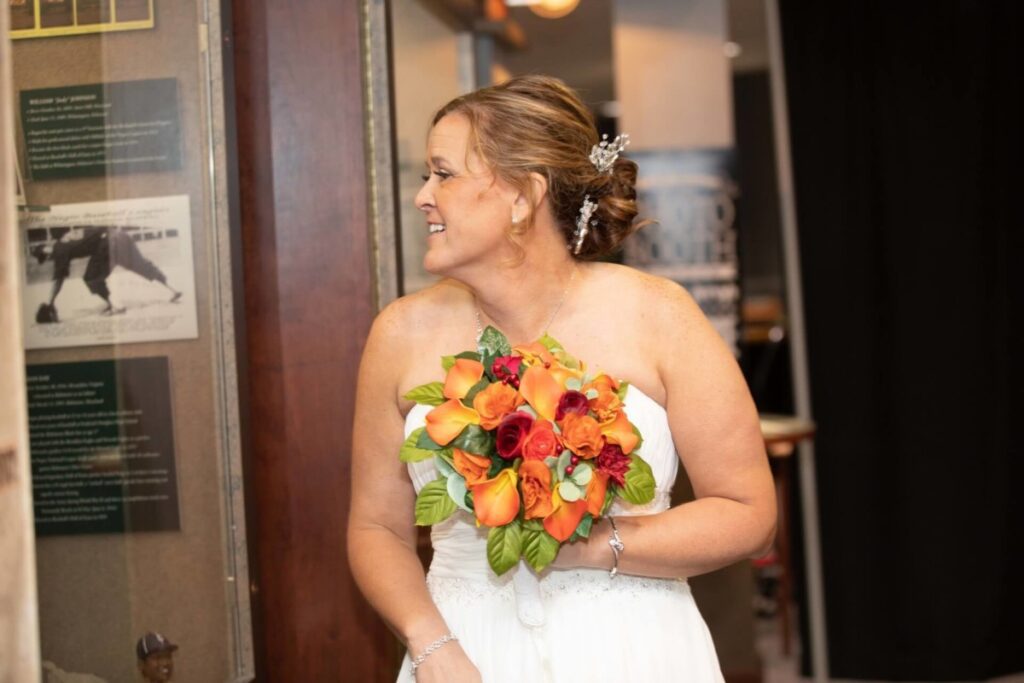 Heather-Scott-Wedding-Low-Res-115 1 - TOMMY GATZ ENTERTAINMENT A bride holding a bouquet of orange and red flowers looks to her right, standing indoors next to a wall with framed photos and text.