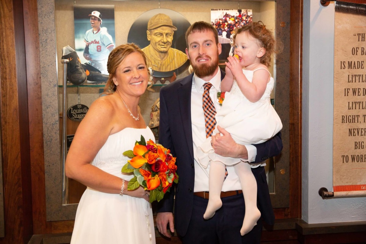 Heather-Scott-Wedding-Low-Res-116 1 - TOMMY GATZ ENTERTAINMENT A bride and groom pose with a young girl in a museum setting, standing in front of sports memorabilia. The bride holds a bouquet, and the girl is holding a small item up to her mouth.