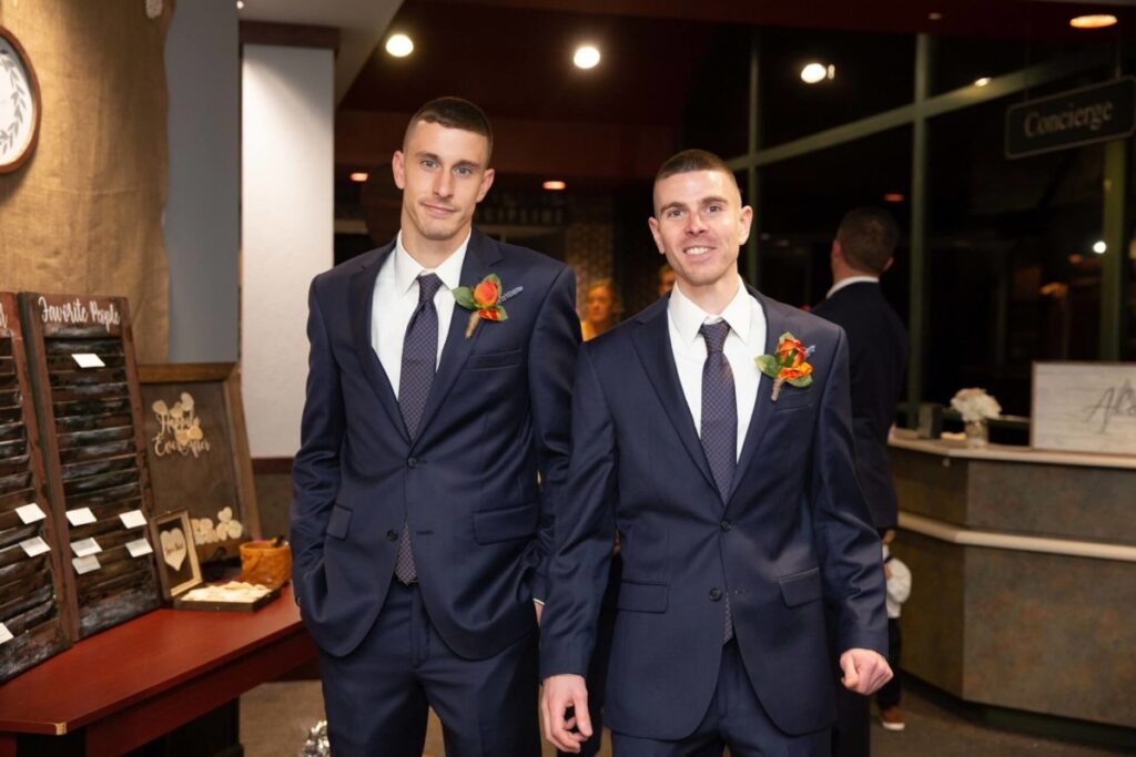 Heather-Scott-Wedding-Low-Res-117 1 - TOMMY GATZ ENTERTAINMENT Two men in matching dark suits with orange boutonnieres stand side by side indoors, with smiles, in a formal setting.