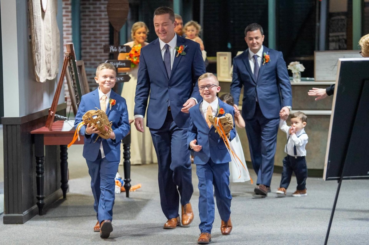 Heather-Scott-Wedding-Low-Res-118 1 - TOMMY GATZ ENTERTAINMENT A group of people in suits, including two boys holding baseball gloves, walk together smiling indoors.