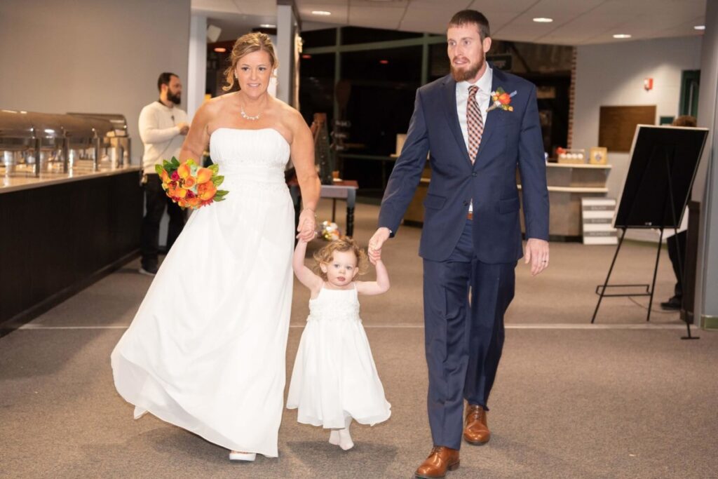 Heather-Scott-Wedding-Low-Res-119 1 - TOMMY GATZ ENTERTAINMENT A couple in formal attire walks with a young girl dressed in white. The woman holds a bouquet of orange flowers.
