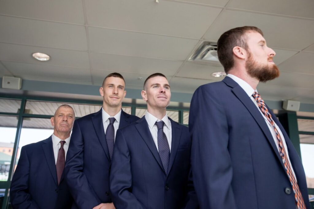 Heather-Scott-Wedding-Low-Res-12 1 - TOMMY GATZ ENTERTAINMENT Four men in suits stand in a row indoors, gazing forward.
