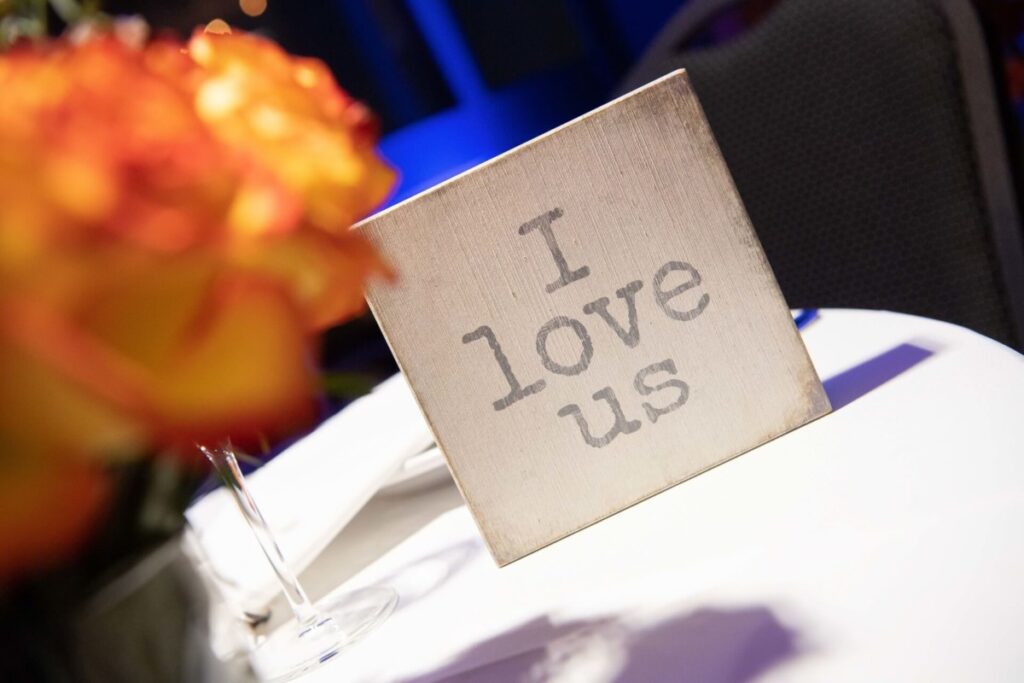 Heather-Scott-Wedding-Low-Res-121 1 - TOMMY GATZ ENTERTAINMENT A small sign on a table reads "I love us," with orange flowers blurred in the foreground.