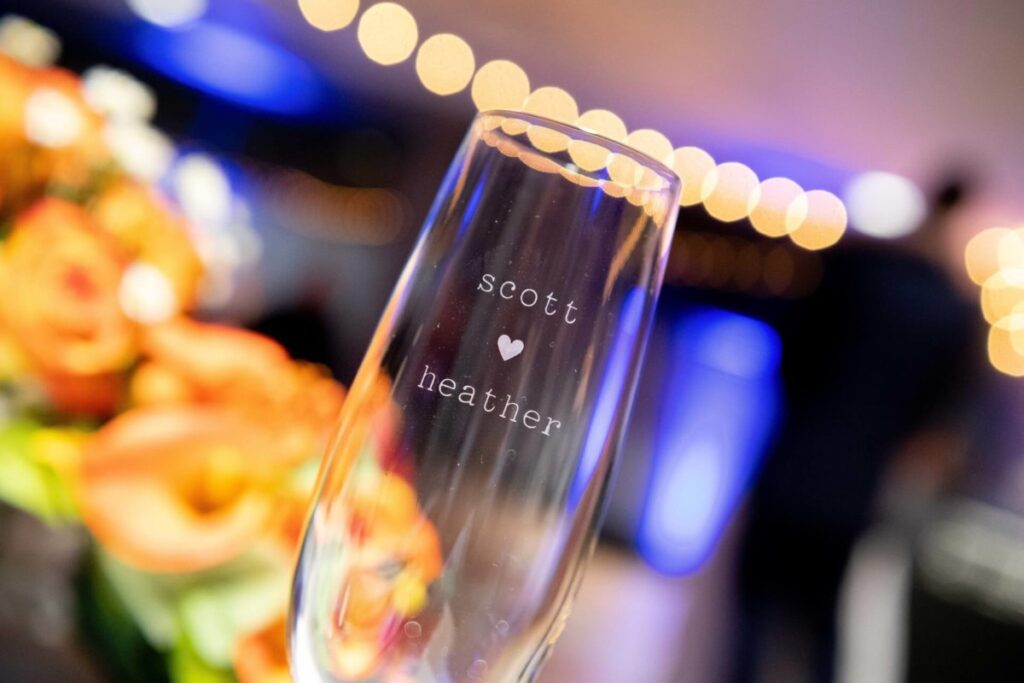 Heather-Scott-Wedding-Low-Res-122 1 - TOMMY GATZ ENTERTAINMENT A champagne flute reads "Scott ♥ Heather," with blurred orange flowers and bokeh lights in the background.