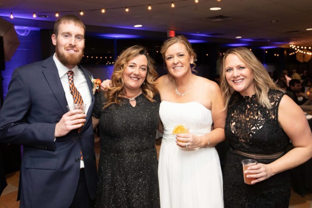Heather-Scott-Wedding-Low-Res-123 1 - TOMMY GATZ ENTERTAINMENT Group of four people smiling at an indoor event, with two holding drinks. The setting has string lights and a lively atmosphere.