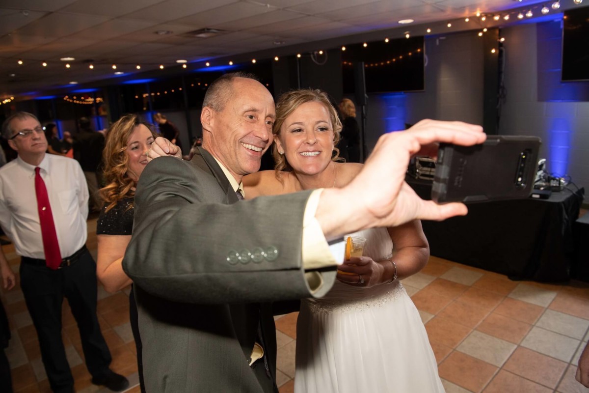 Heather-Scott-Wedding-Low-Res-125 1 - TOMMY GATZ ENTERTAINMENT A man and woman take a selfie at an indoor event with people around them.