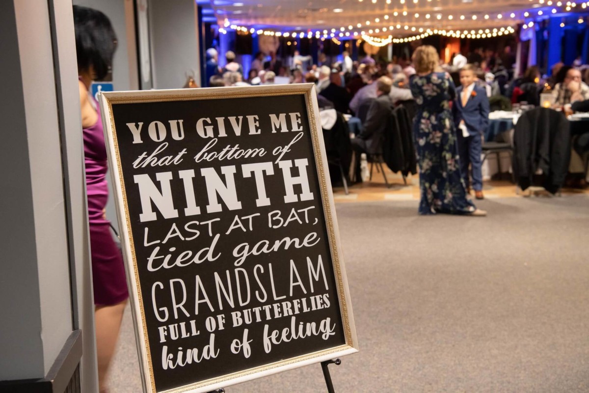Heather-Scott-Wedding-Low-Res-129 1 - TOMMY GATZ ENTERTAINMENT Sign with text about a baseball game feeling in the foreground; people socializing in a decorated venue in the background.