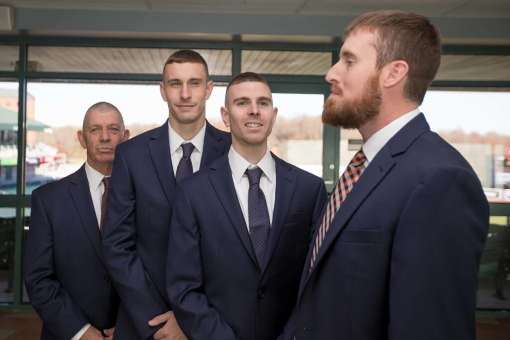 Heather-Scott-Wedding-Low-Res-13 1 - TOMMY GATZ ENTERTAINMENT Four men in suits standing in line indoors, with a windowed background.