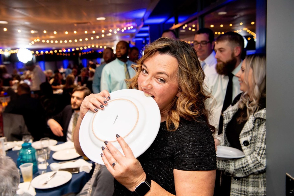 Heather-Scott-Wedding-Low-Res-132 1 - TOMMY GATZ ENTERTAINMENT A woman playfully pretends to bite a plate at a crowded event with string lights overhead.