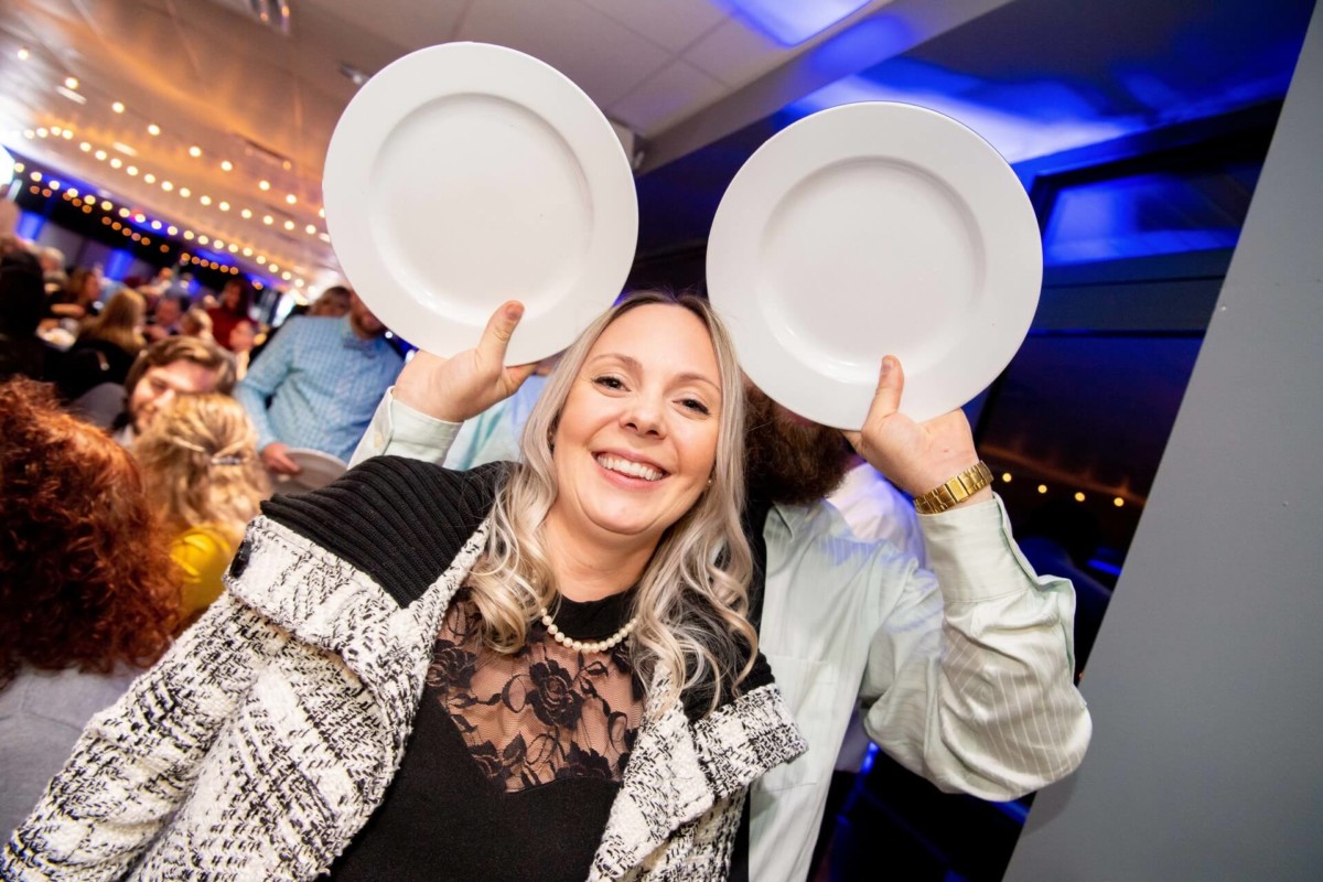 Heather-Scott-Wedding-Low-Res-133 1 - TOMMY GATZ ENTERTAINMENT A woman smiles at the camera while someone behind her holds two plates near her head, creating a humorous effect resembling ears.