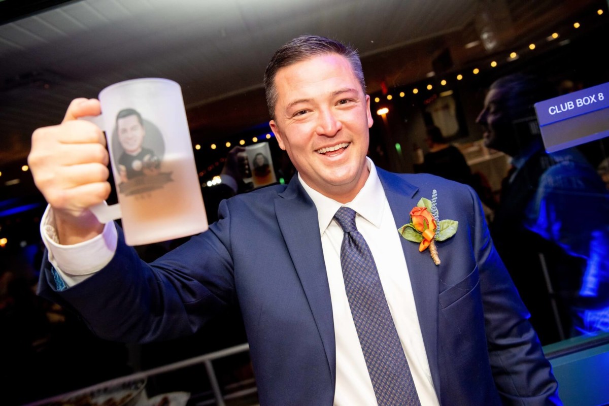Heather-Scott-Wedding-Low-Res-134 1 - TOMMY GATZ ENTERTAINMENT A man in a suit smiles and raises a frosted glass mug with a picture on it. He has a boutonniere on his lapel.