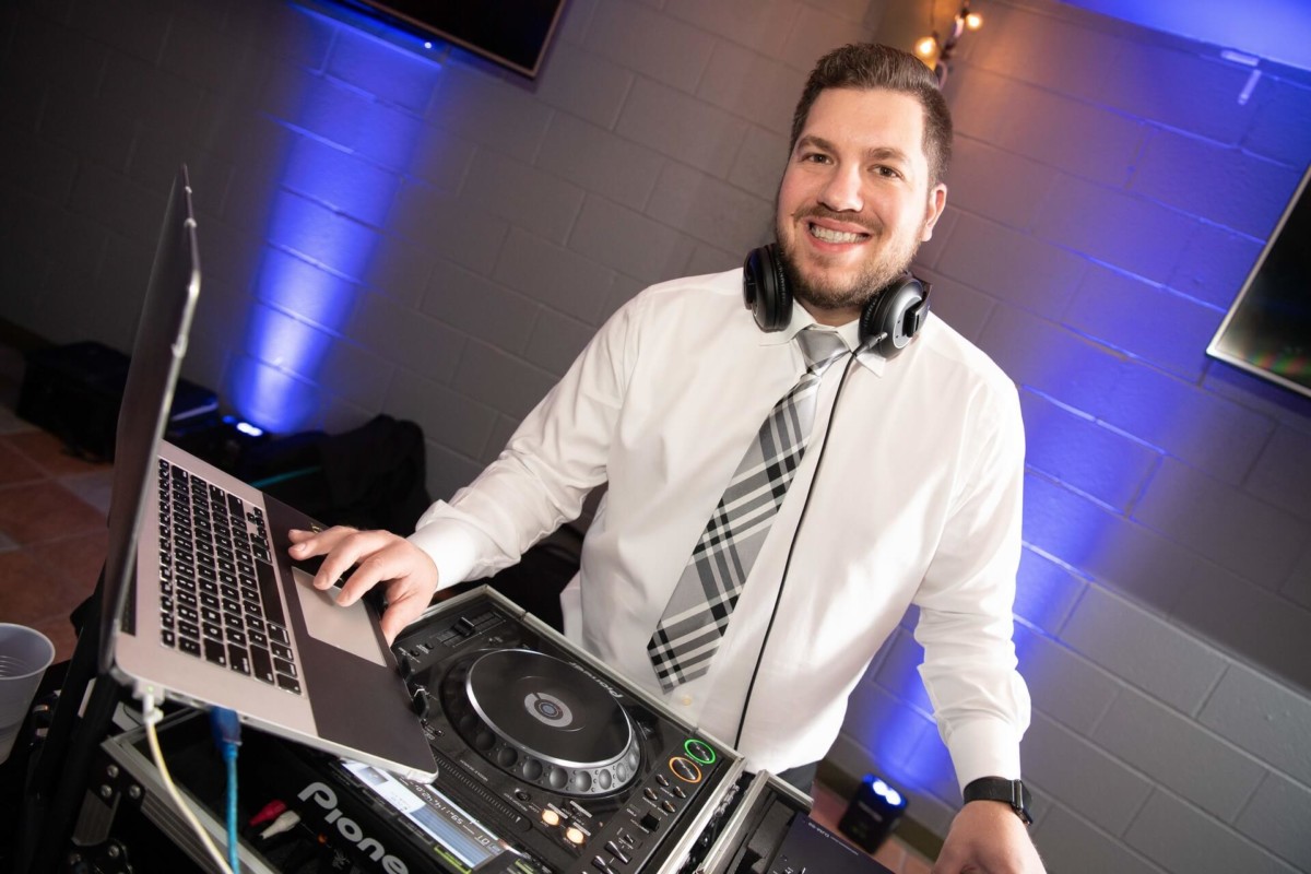 Heather-Scott-Wedding-Low-Res-138 1 - TOMMY GATZ ENTERTAINMENT A man in a white shirt and tie is DJing at an event. He stands behind a console with headphones around his neck and a laptop in front of him.