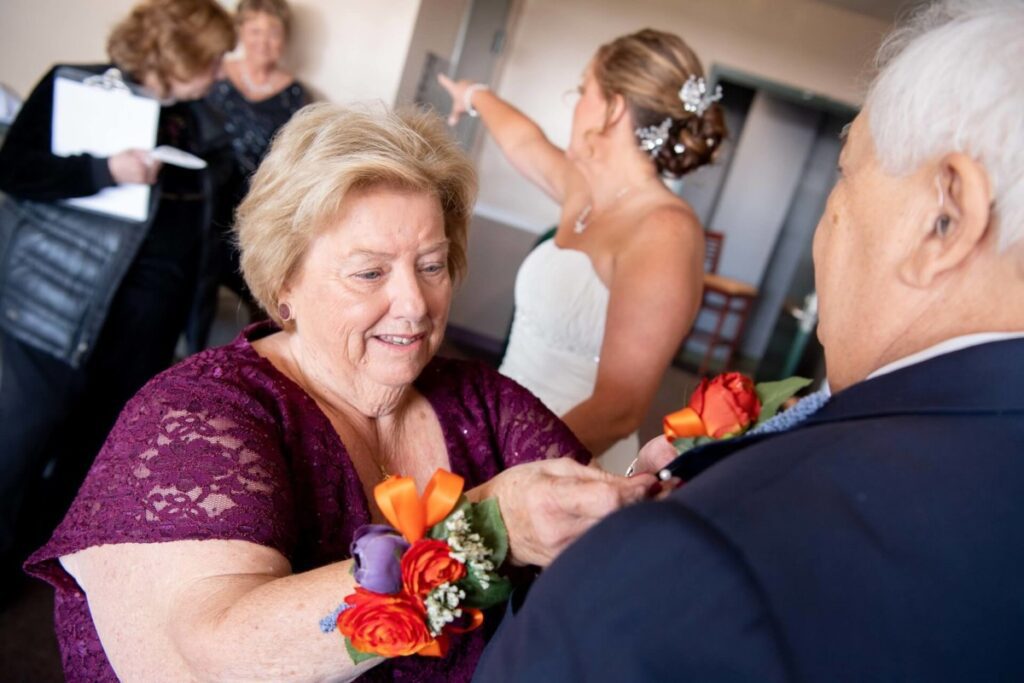 Heather-Scott-Wedding-Low-Res-14 1 - TOMMY GATZ ENTERTAINMENT An elderly woman in a purple dress pins a boutonnière on an elderly man's suit. A bride and two other women are in the background.
