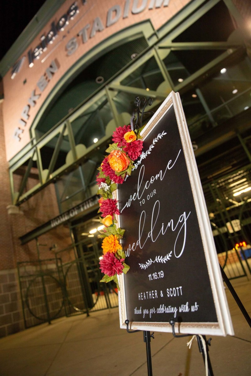 Heather-Scott-Wedding-Low-Res-141 1 - TOMMY GATZ ENTERTAINMENT Wedding welcome sign with flowers in front of a building labeled Ripken Stadium.