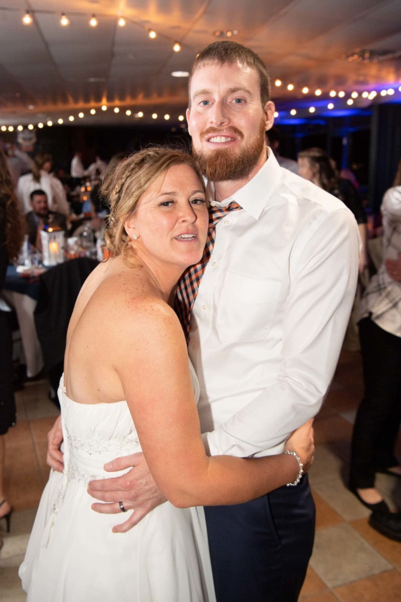 Heather-Scott-Wedding-Low-Res-142 1 - TOMMY GATZ ENTERTAINMENT A couple embraces at a festive indoor event, surrounded by string lights, tables, and guests.
