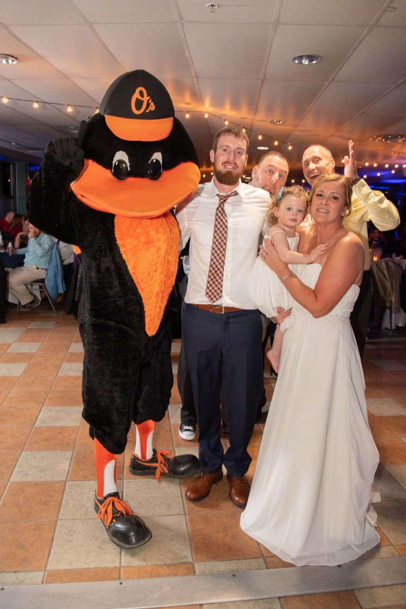 Heather-Scott-Wedding-Low-Res-144 1 - TOMMY GATZ ENTERTAINMENT A group posing with a person in a Baltimore Orioles mascot costume at a celebration. A bride holds a child, while two other men stand smiling.