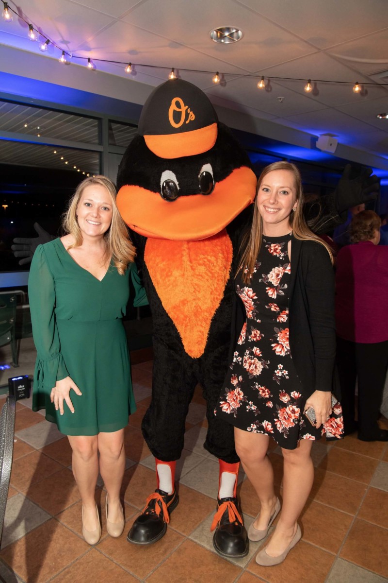 Heather-Scott-Wedding-Low-Res-145 1 - TOMMY GATZ ENTERTAINMENT Two women pose with a person in an Orioles bird mascot costume indoors, under ceiling lights.