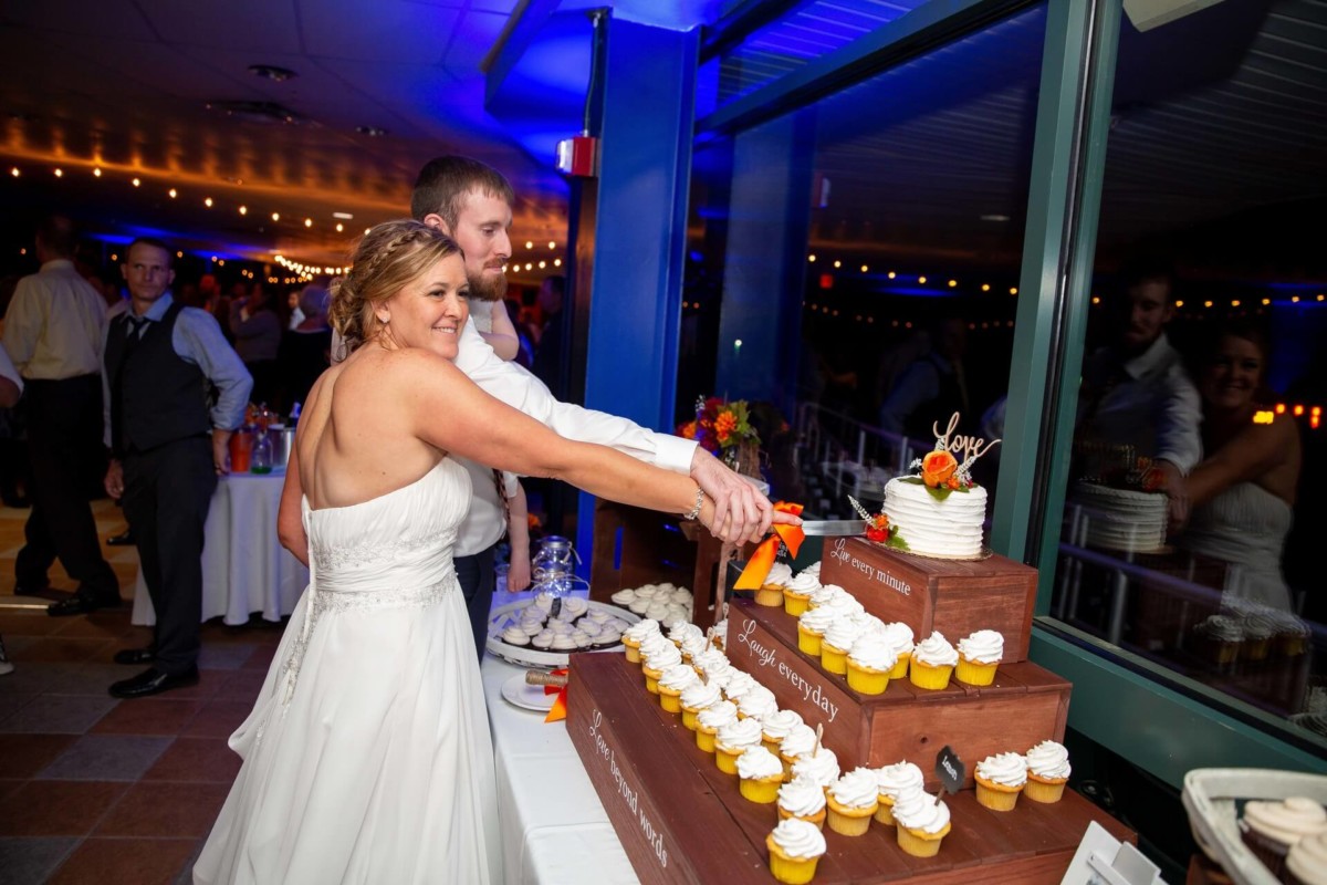 Heather-Scott-Wedding-Low-Res-146 1 - TOMMY GATZ ENTERTAINMENT A bride and groom cut into a tiered wedding cake surrounded by cupcakes, with a small group of guests in the background.