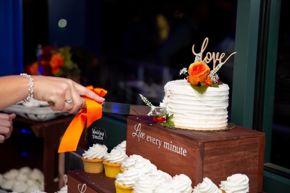 Heather-Scott-Wedding-Low-Res-147 1 - TOMMY GATZ ENTERTAINMENT A person cuts a small white cake with an orange ribbon, adorned with a rose and a "love" topper. Cupcakes below have "Live every minute" displayed on wooden stands.