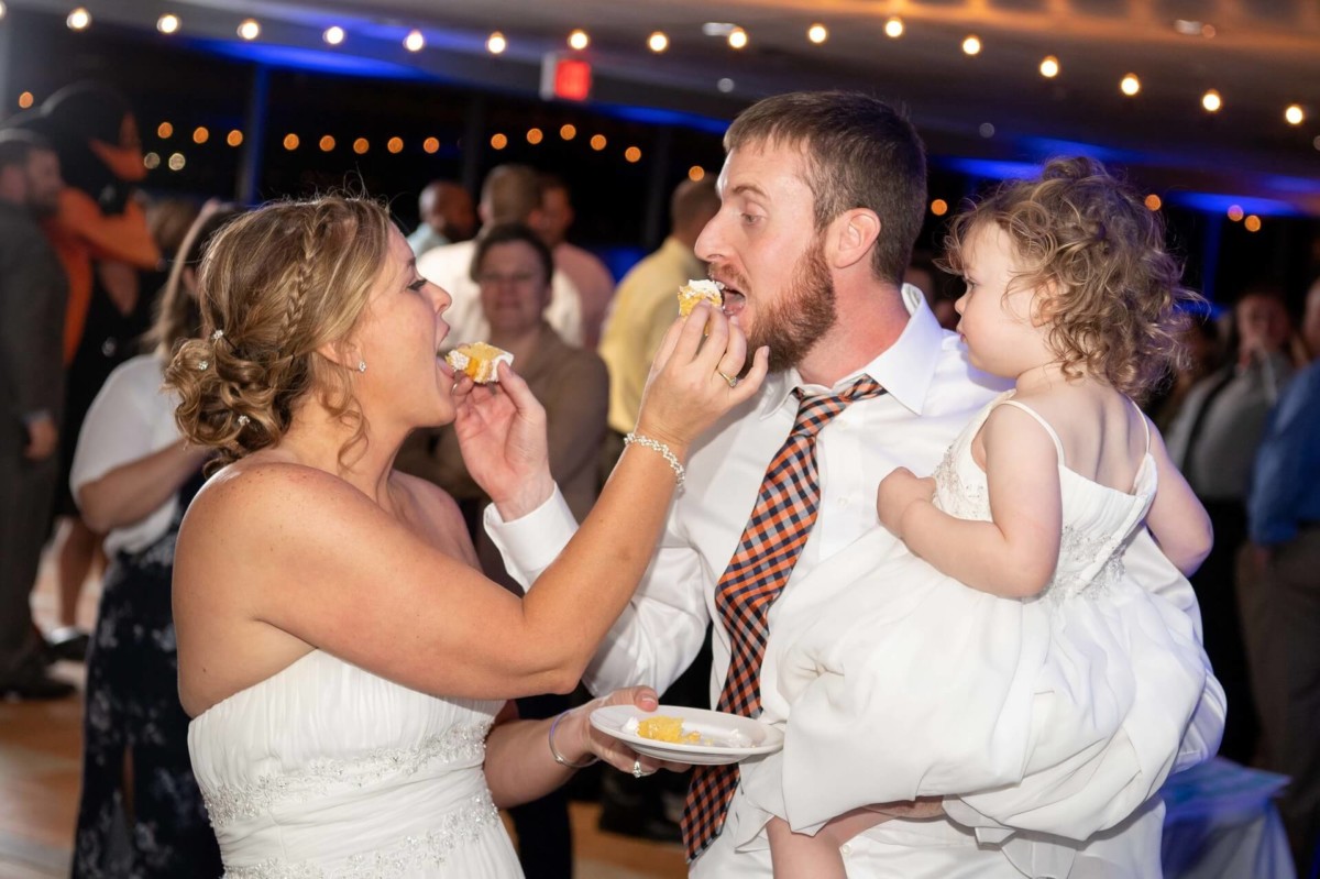 Heather-Scott-Wedding-Low-Res-148 1 - TOMMY GATZ ENTERTAINMENT A bride and groom feed each other cake at a wedding reception. The groom holds a young child who looks on. Guests are visible in the background.