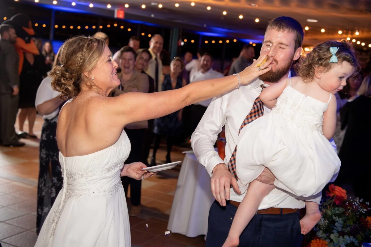 Heather-Scott-Wedding-Low-Res-149 1 - TOMMY GATZ ENTERTAINMENT A bride playfully smears cake on a man's face while he holds a child. Guests in the background smile and laugh.