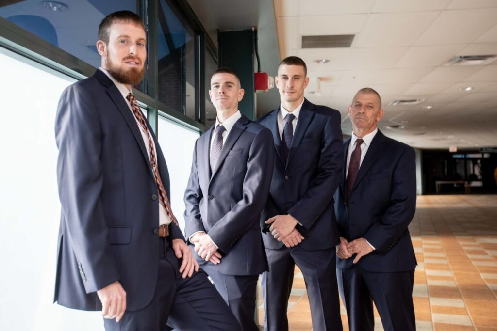 Heather-Scott-Wedding-Low-Res-15 1 - TOMMY GATZ ENTERTAINMENT Four men in suits stand together in a well-lit corridor, posing and looking at the camera.