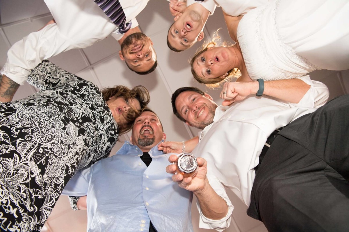 Heather-Scott-Wedding-Low-Res-150 1 - TOMMY GATZ ENTERTAINMENT A group of people in formal wear are looking down, smiling and surprised, with one person holding a small object toward the camera.
