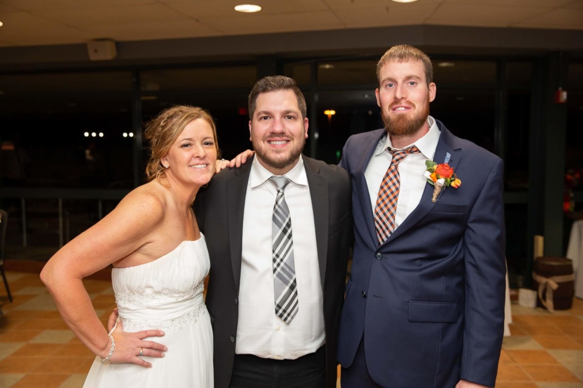 Heather-Scott-Wedding-Low-Res-152 1 - TOMMY GATZ ENTERTAINMENT Three people pose for a photo at an indoor event; the woman on the left is wearing a white dress, and the two men are in suits.
