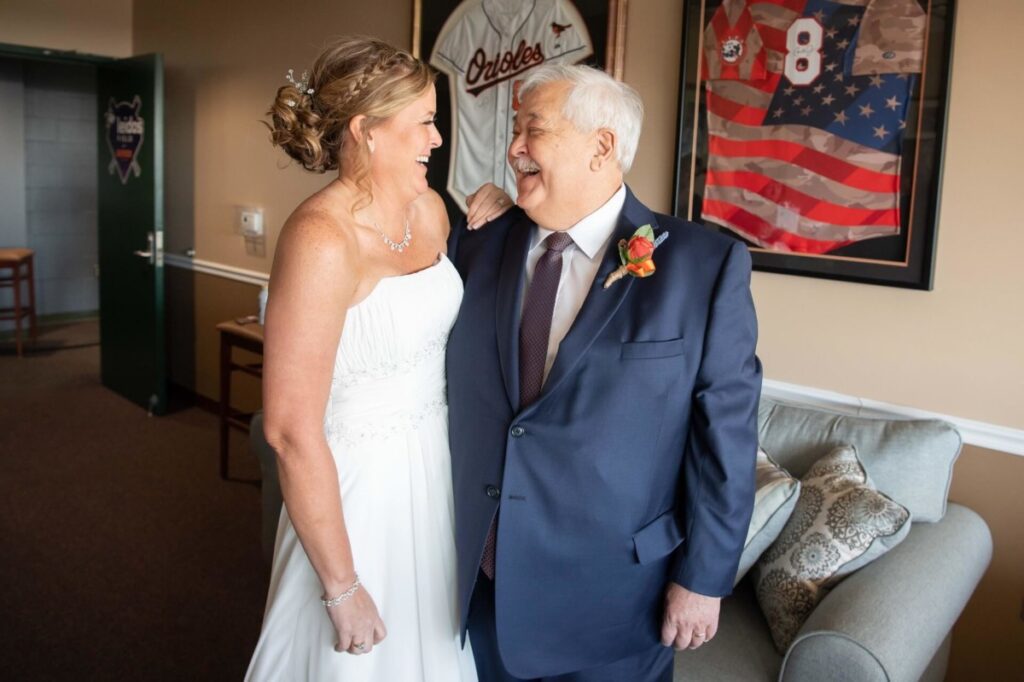 Heather-Scott-Wedding-Low-Res-16 1 - TOMMY GATZ ENTERTAINMENT Bride in a white dress and older man in a suit smile at each other in a room with sports memorabilia on the walls.