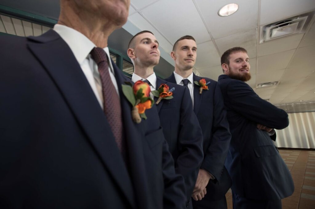 Heather-Scott-Wedding-Low-Res-17 1 - TOMMY GATZ ENTERTAINMENT Four men in suits with boutonnieres stand indoors; three are facing forward, and one has arms crossed and is looking to the side.