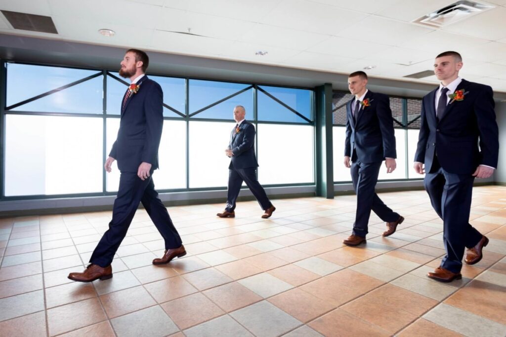 Heather-Scott-Wedding-Low-Res-18 1 - TOMMY GATZ ENTERTAINMENT Four men in suits walk in a line across a tiled floor in a bright room with large windows.